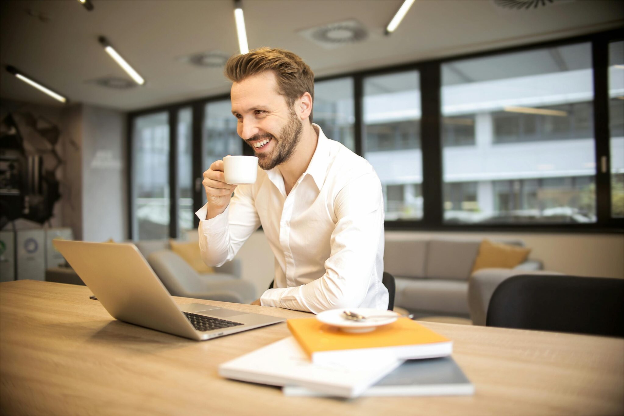 Man enjoying a coffee break while working in a modern office with laptop and books.